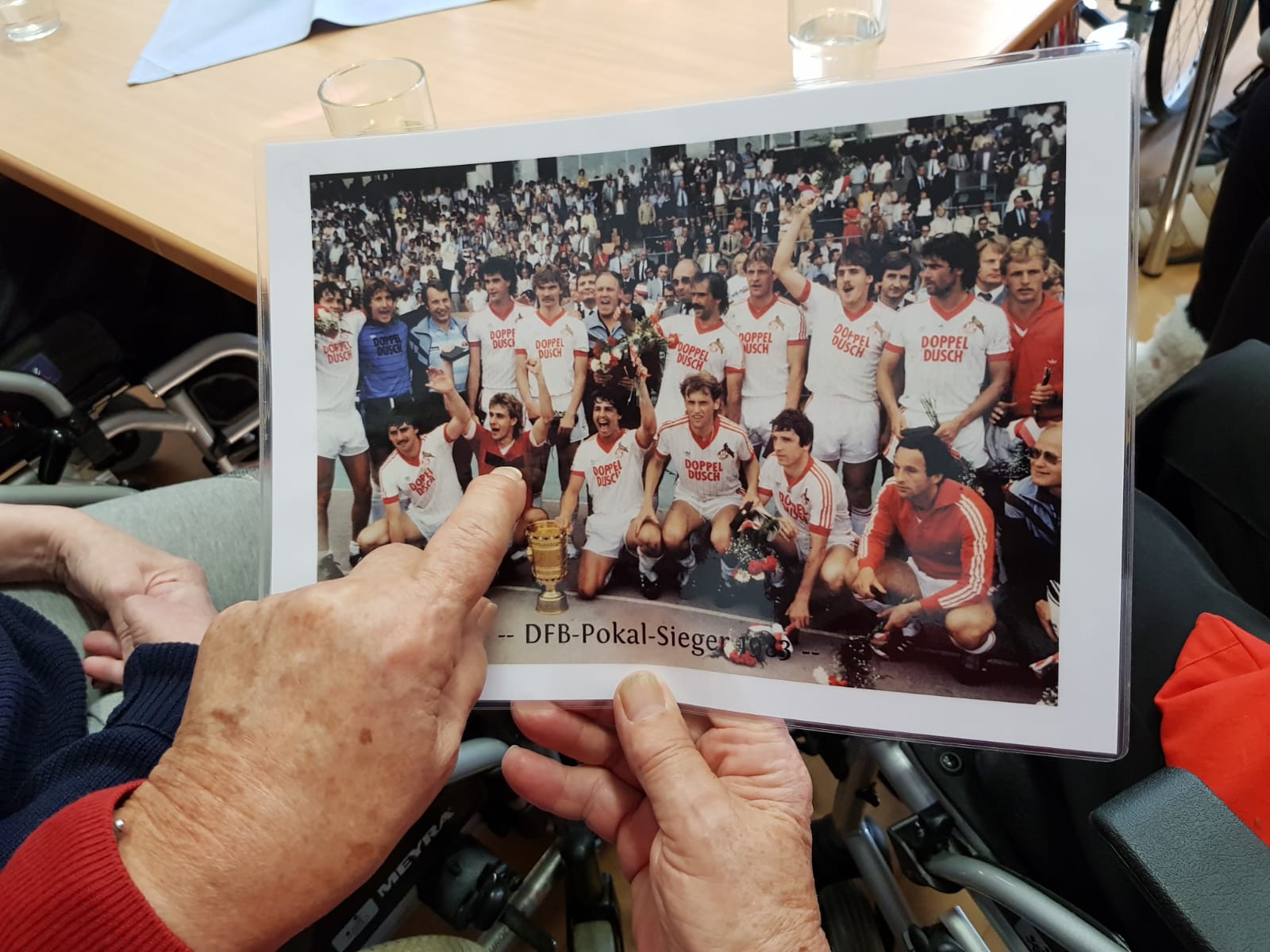 Zwei Hände halten ein älteres Mannschaftsbild des 1. FC Köln. Die linke Hand zeigt mit dem Finger auf einen Spieler.