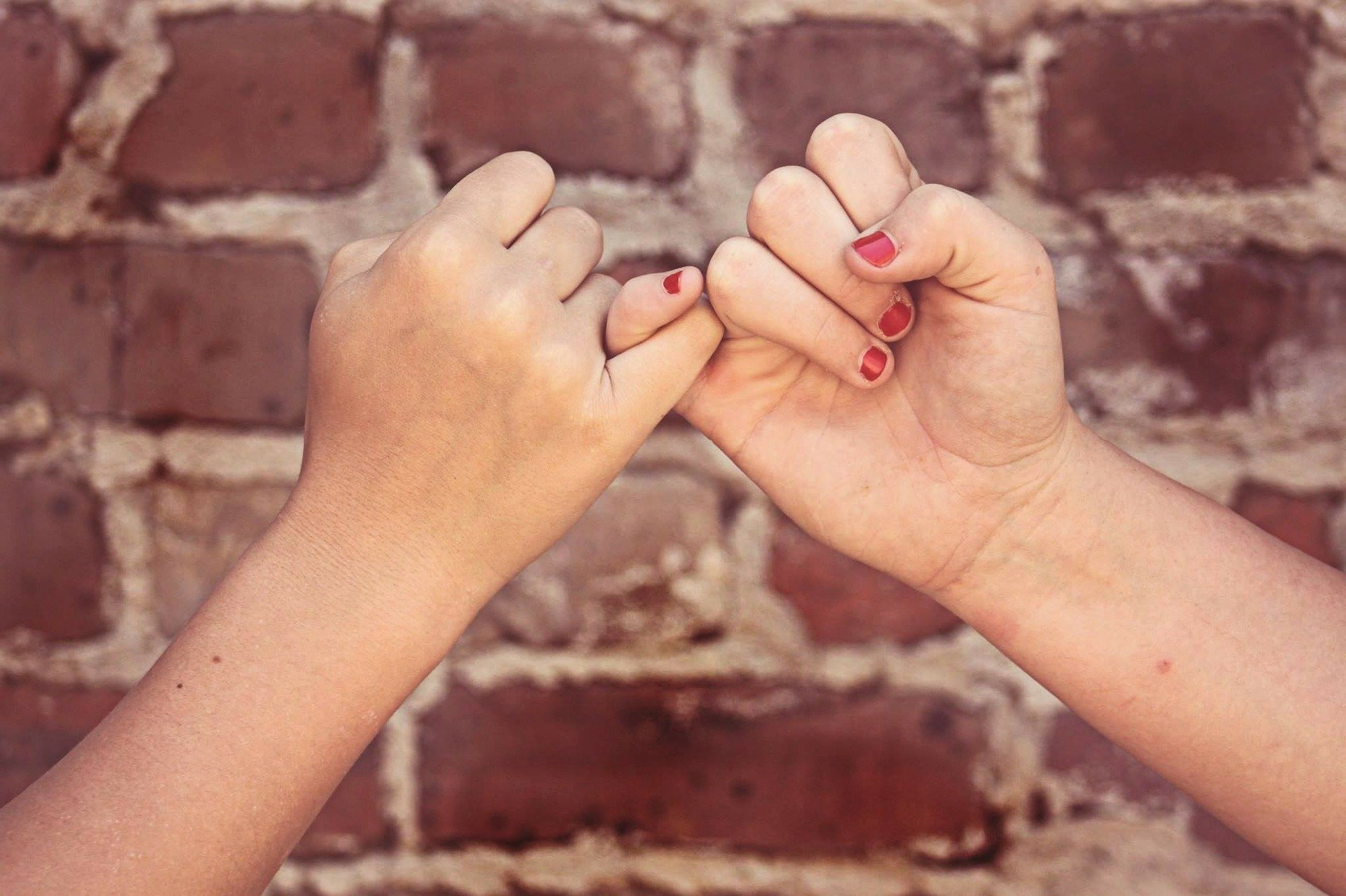 Zwei Hände deren kleine Finger ineinander verhakt sind. Im Hintergrund eine rote Ziegelsteinmauer.