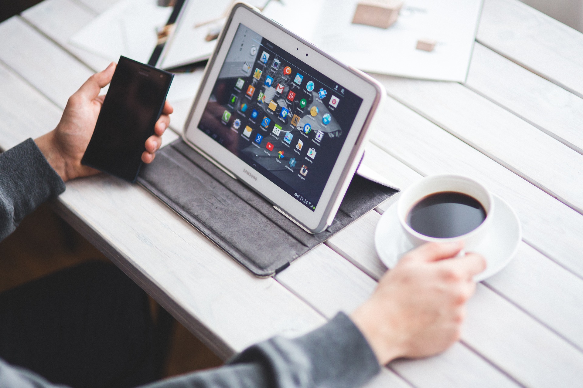 Ein Mensch sitzt am Tisch, man sieht nur die Arme. In der linken Hand ein Smartphone, in der rechten eine Tasse Kaffee. Mittig steht auf einem Ständer ein weißes Tablet. Auf dem Display sieht man die Icons der unterschiedlichen Apps.