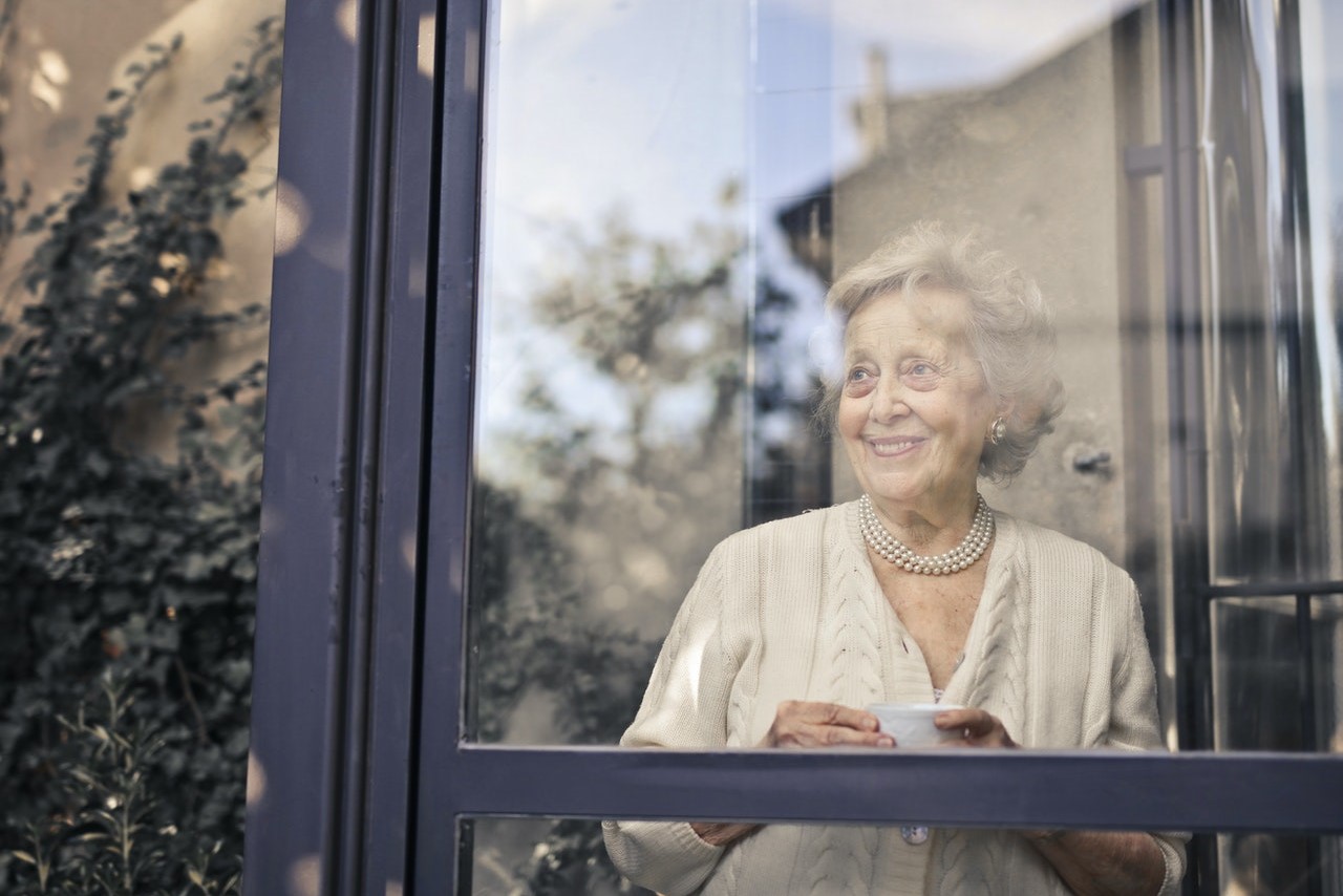 Eine Seniorin steht lächelnd hinter einem Fenster. Sie hat eine Tasse in der Hand.