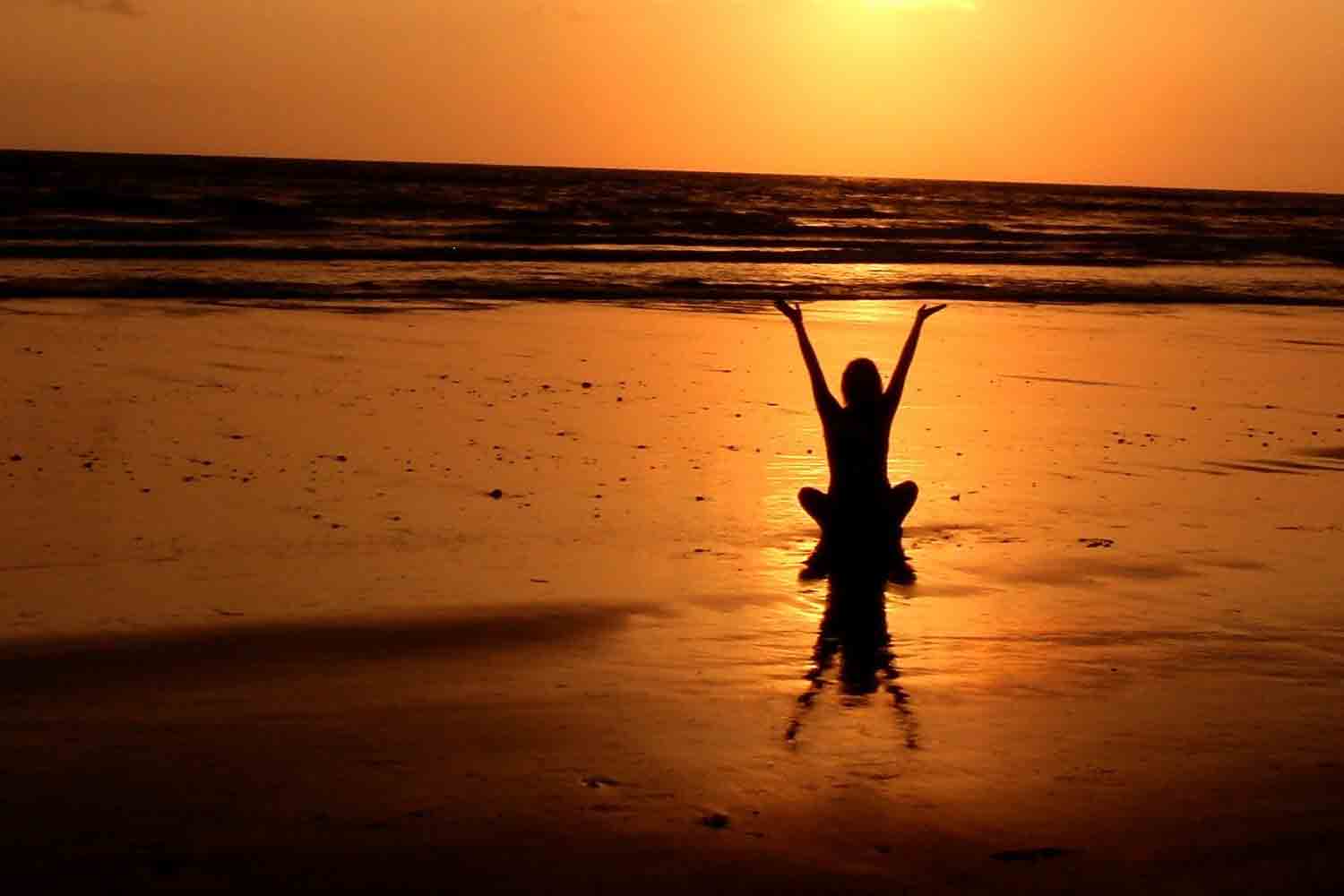 Eine Person macht Yoga am Strand mit dem Blick in Richtung Sonnenuntergang.