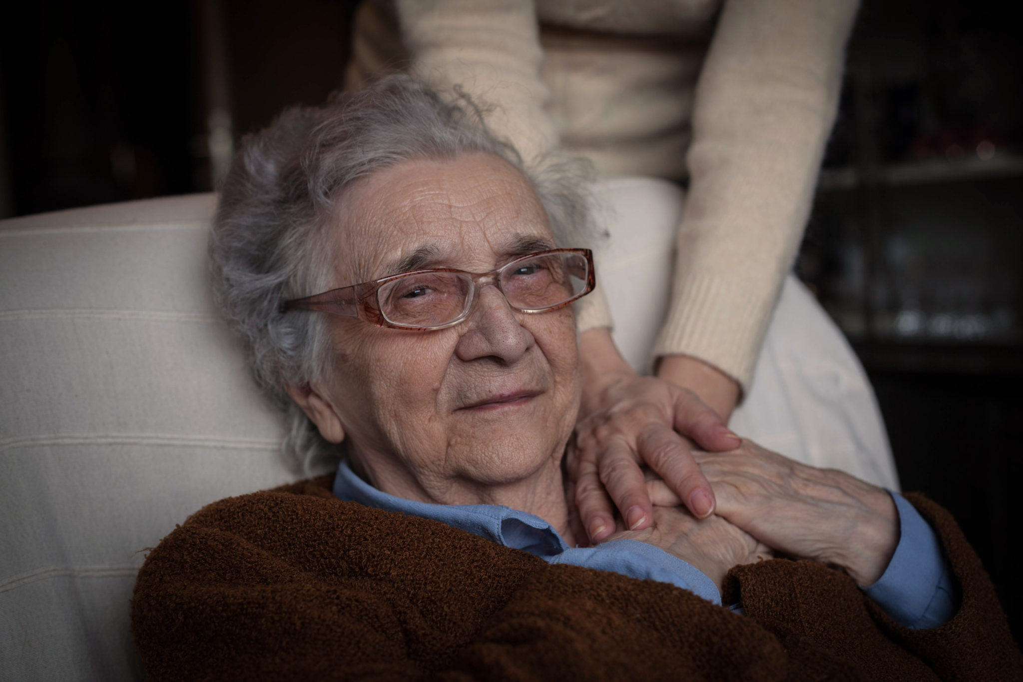 Eine Seniorin sitzt auf einem Sessel und hält die Hand einer jüngeren Frau.