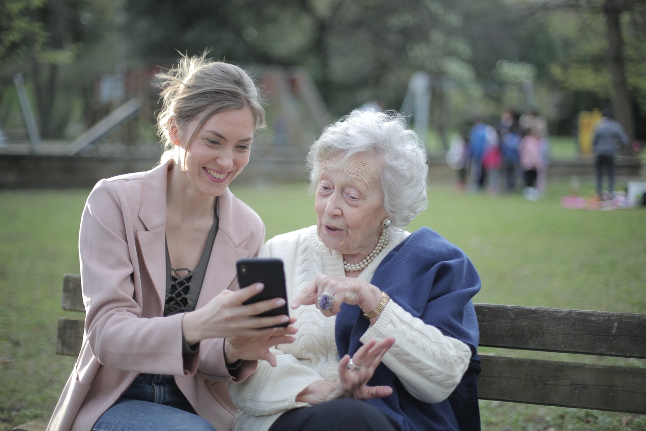 Eine junge Frau und eine Senioren sitzen auf einer Bank in einem Park und bedienen gemeinsam ein Handy.