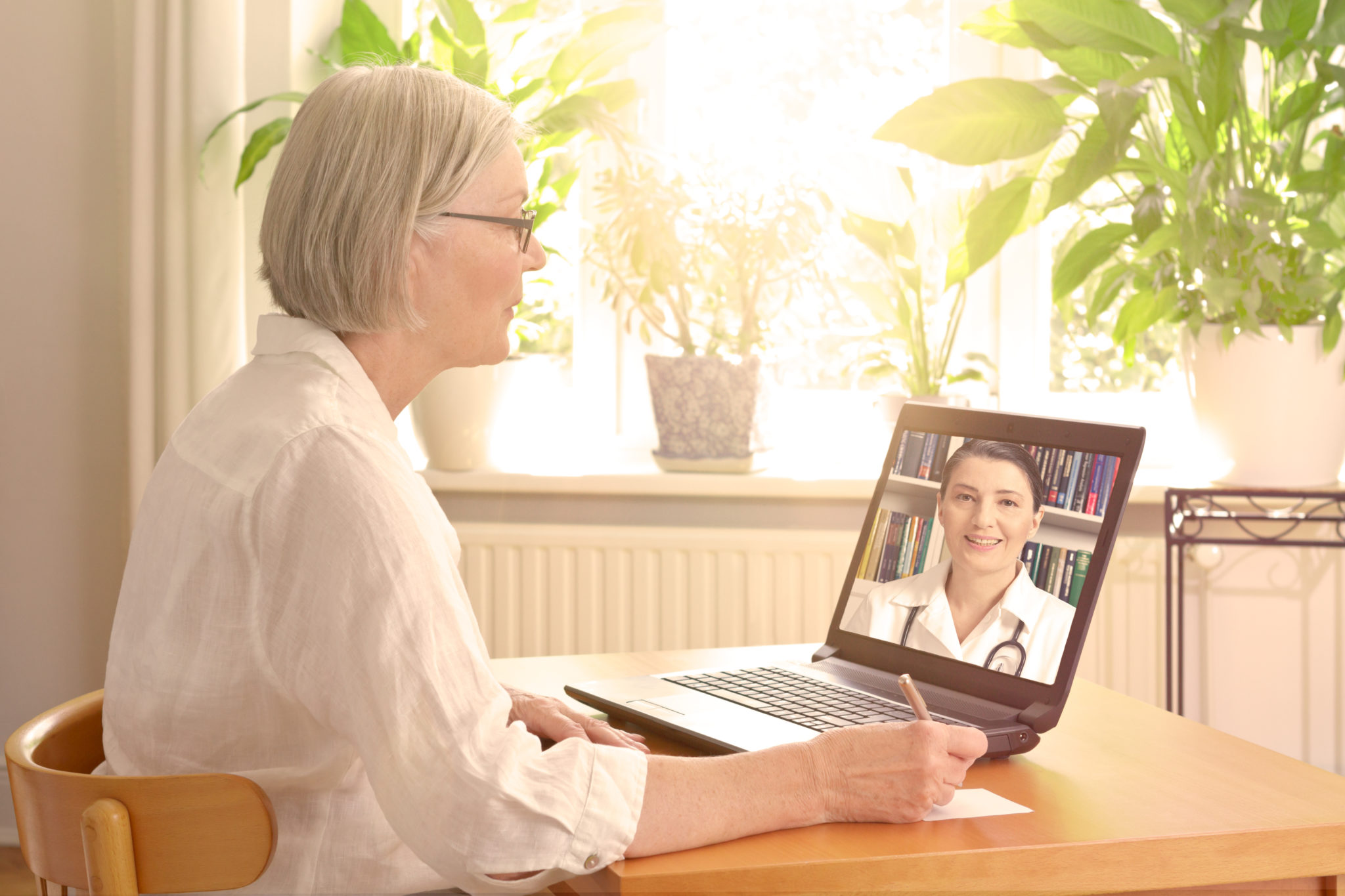 Eine Seniorin sitzt am Tisch in ihrer Wohnung und nimmt an einer Onlinekonferenz teil.