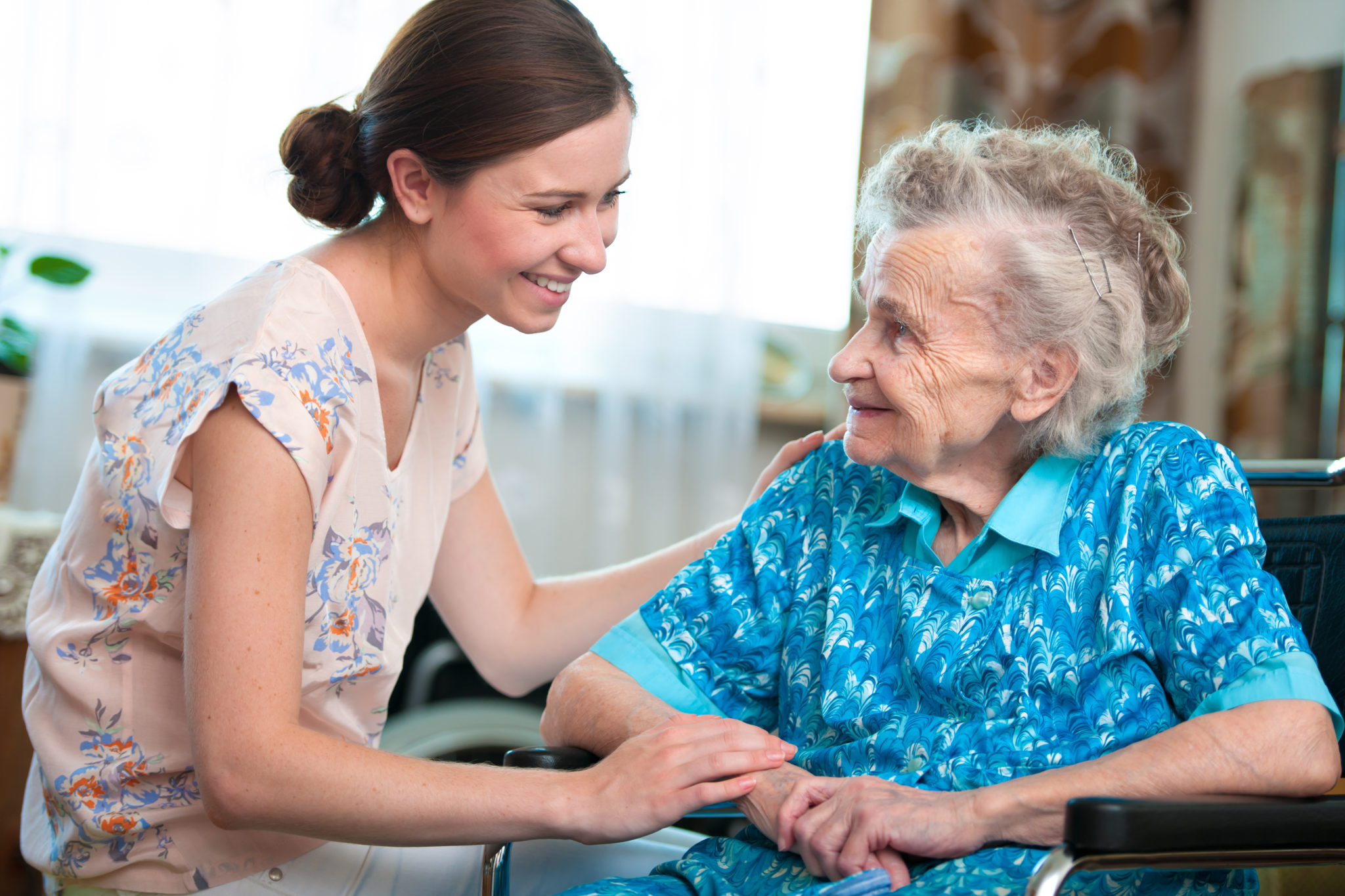 Zu sehen sind eine pflegebedürftige Seniorin und ihre Enkelin. Beide lächeln sich an.