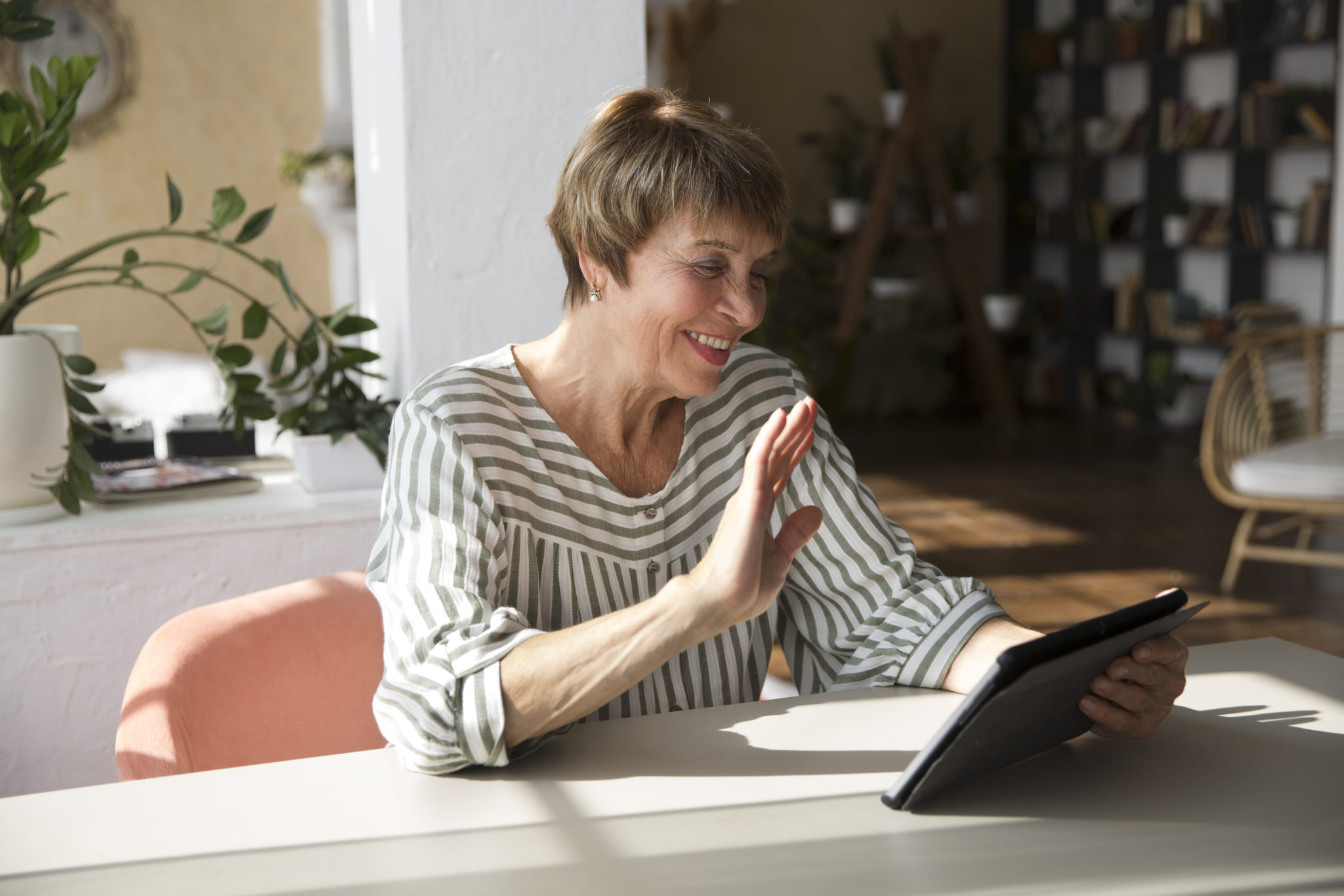 Eine Frau sitzt an einem Tisch und nutzt ein Tablet für einen Videoanruf.. Sie winkt in die Kamera.