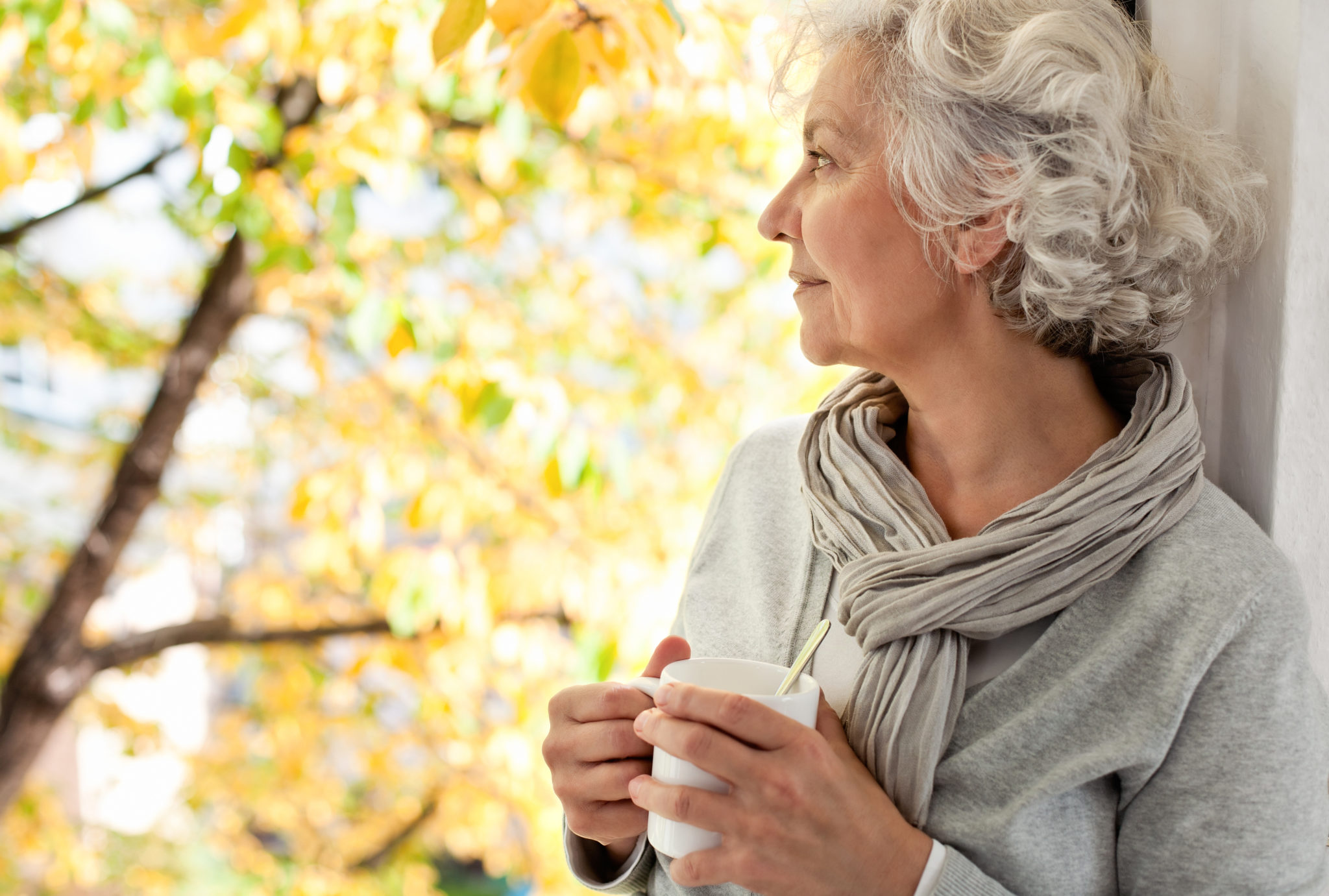 Eine Seniorin schaut alleine mit einer Tasse Kaffe aus dem Fenster. Sie macht den Eindruck, dass sie einsam ist.