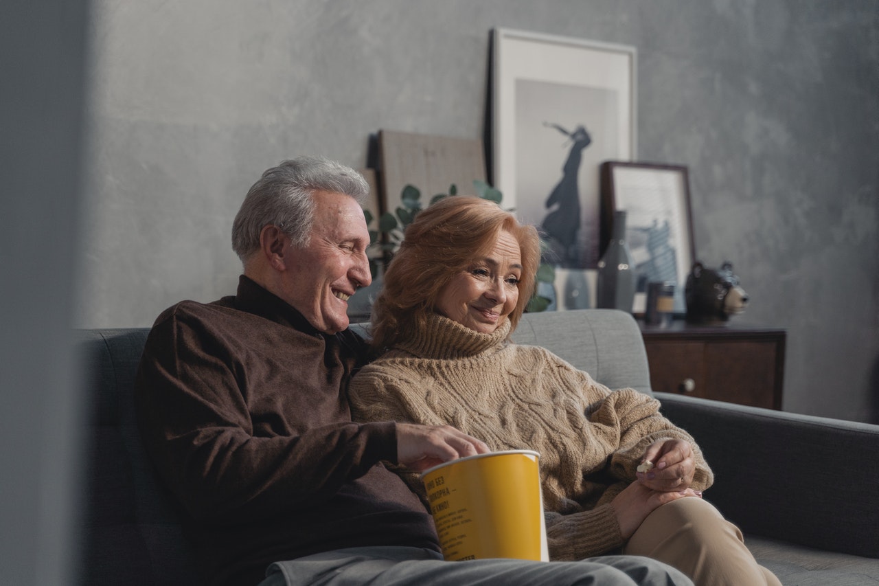 Zwei Senior*innen sitzen auf einer Couch und essen Popcorn. Sie schauen einen FIlm.