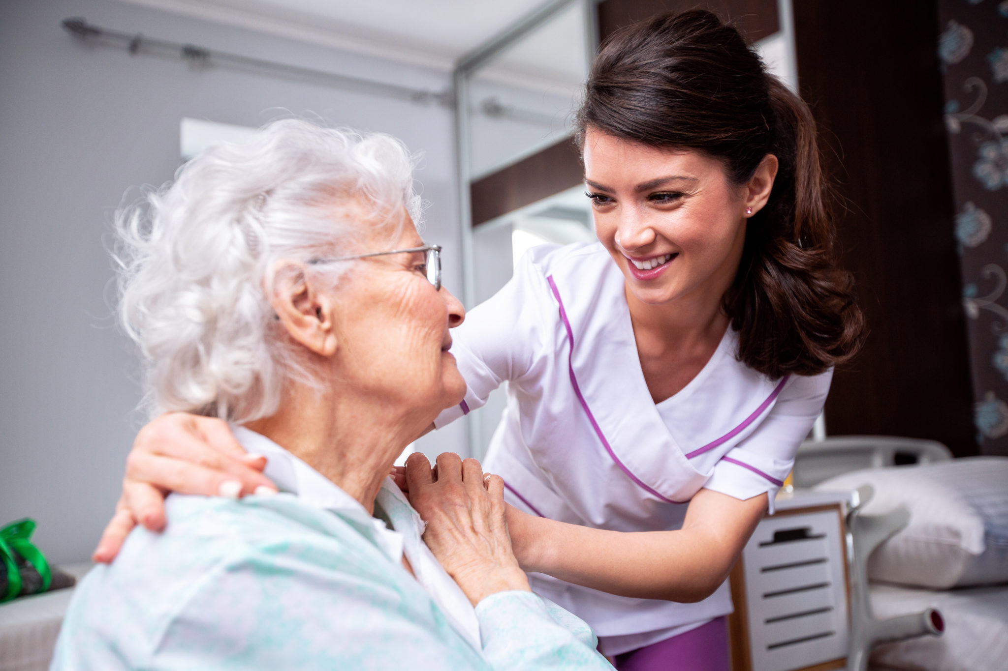 Die Mitarbeiterin eines ambulanten Pflegedienstes lächelt eine Seniorin an, während die ihren Arm um sie legt.