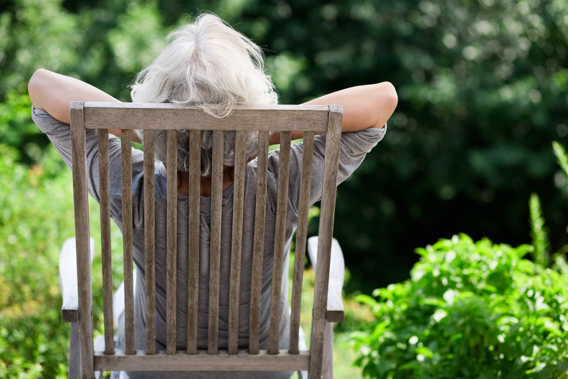 Eine Seniorin lehtn sich bequem auf einem Stuhl zurück und blick in den Garten.