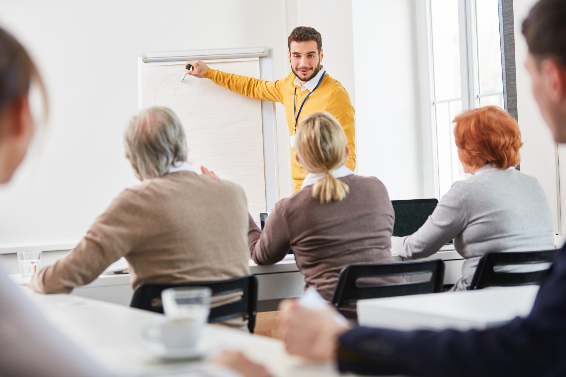 Foto aus einem Seminar. Der Dozent zeigt auf das Flipchart und eine Teilnehmering stellt eine Frage.