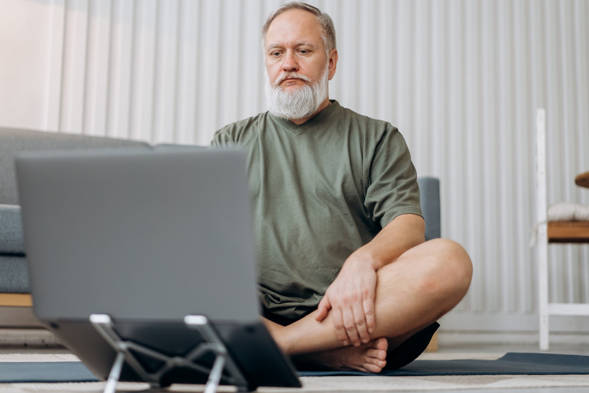 Ein Mann sitzt im Schneidersitz auf einer Yoga-Matte. Vor ihm ist ein Laptop aufgebaut.