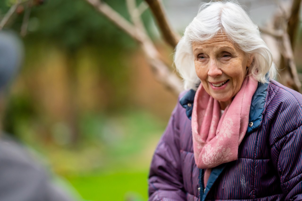 Eine Seniorin ist im Garten und lächtelt in die Kamera.