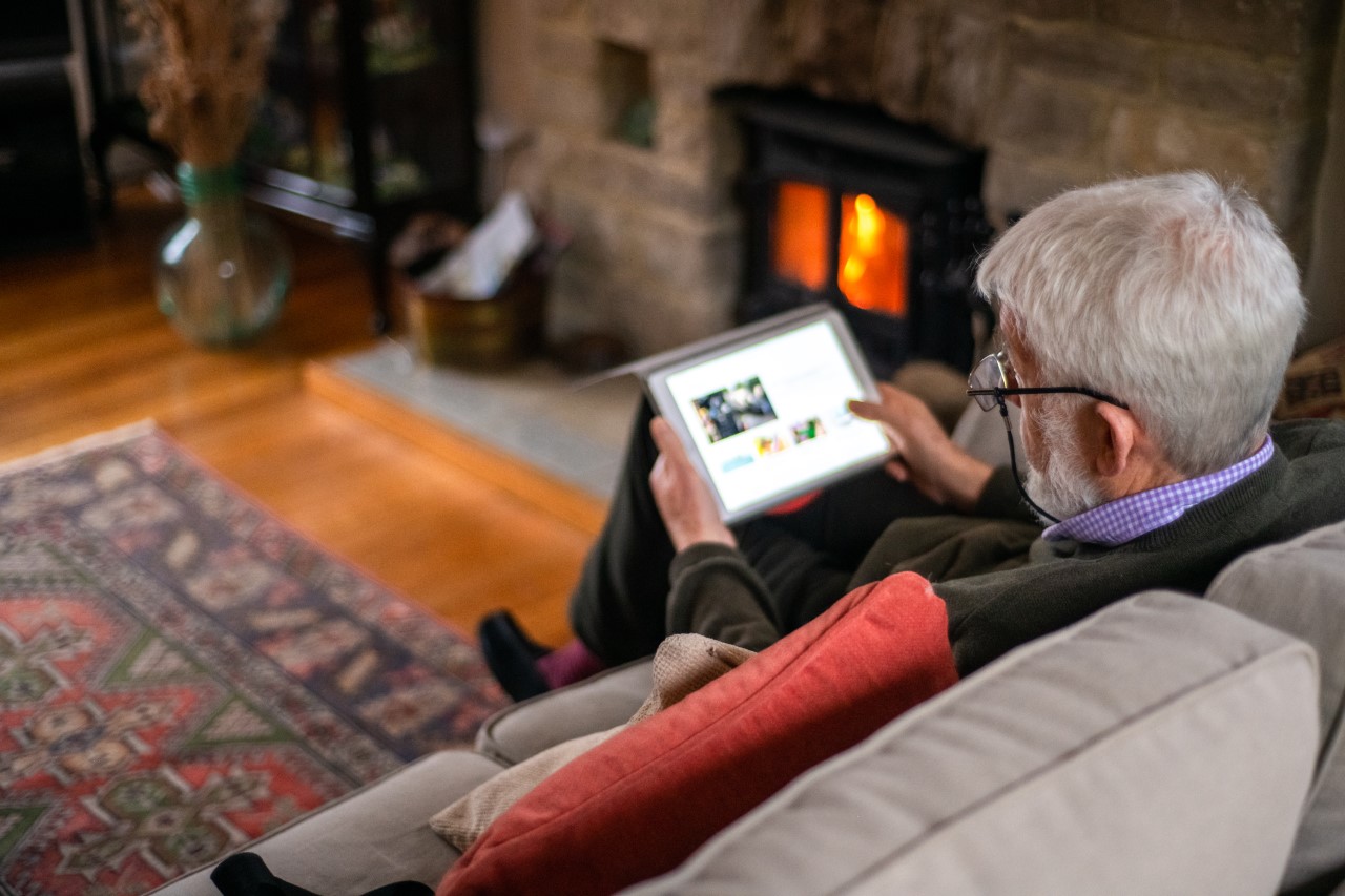 Ein Senior sitzt im Wohnzimmier auf der Couch und bedient ein Tablet.