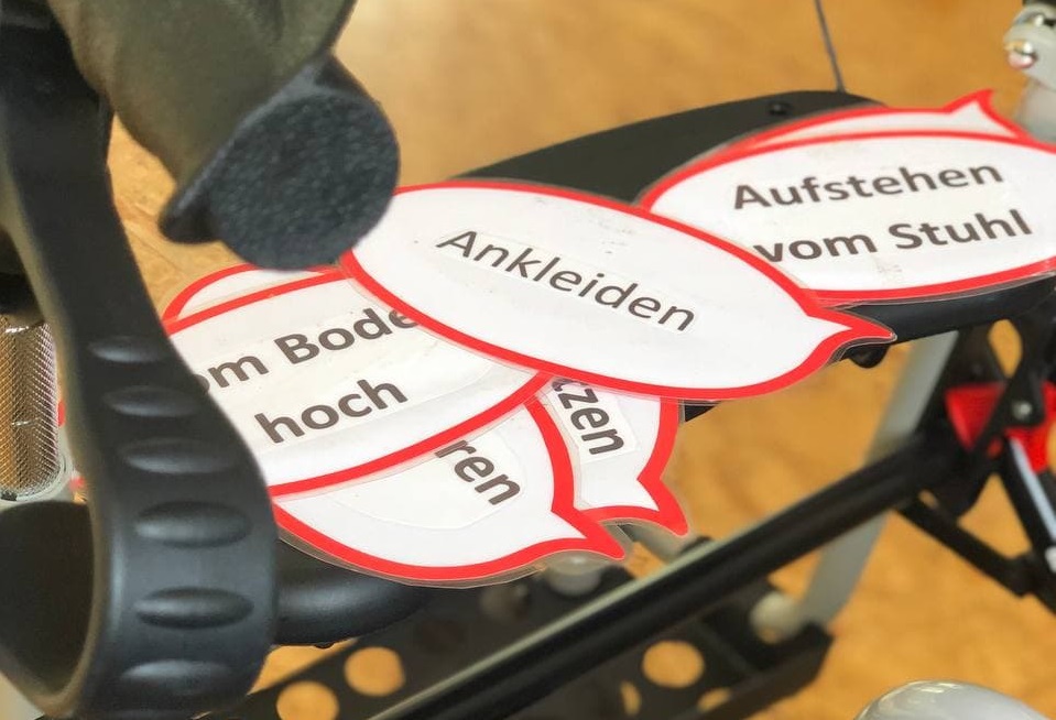 Symbolbild eines Rollators, darauf liegen ausgedruckte Sprechblasen mit texten wie "Ankleiden", "Vom Boden hoch", "Aufstehen vom Stuhl"