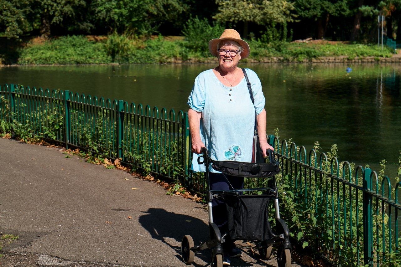 Eine Seniorin geht mit ihrem Rollator im Park spazieren und lacht.