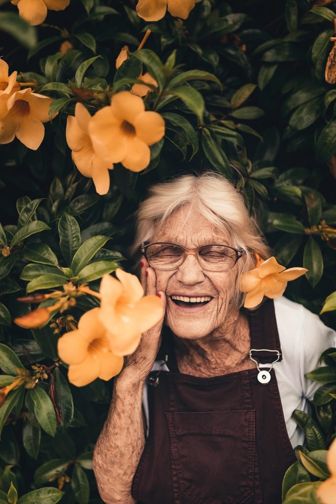 Alte Frau steht vor Busch mit vielen Blüten.