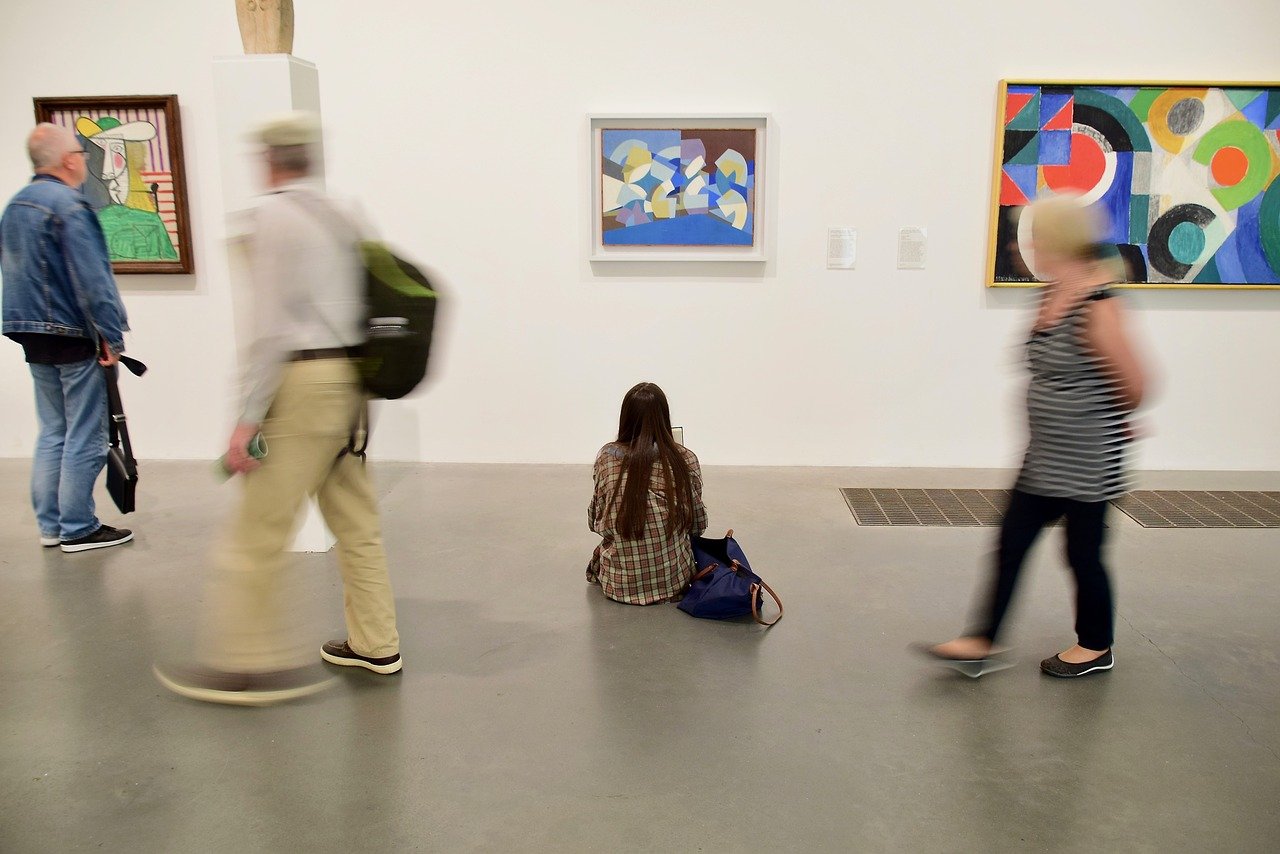 Das Bild zeigt Menschen in einem Museum vor drei Bildern. Eine der Personen sitzt auf dem Boden und betrachtet das Bild in der Mitte.