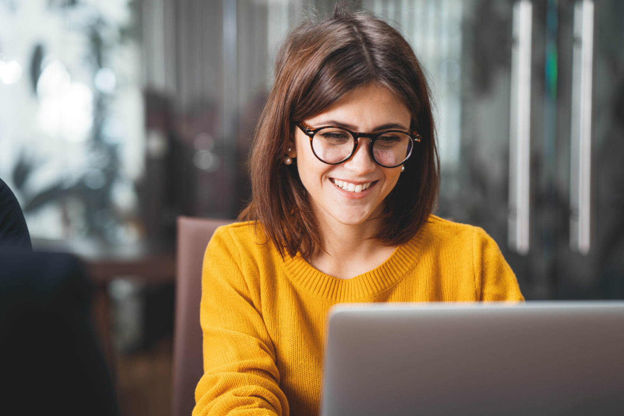 Ein Portät einer glücklichen Buisness-Frau, die Brille trägt an ihrem Arbeitsplatz. Die junge Frau sitzt vor ihrem modernen Laptop und schaut auf den Bildschrim.