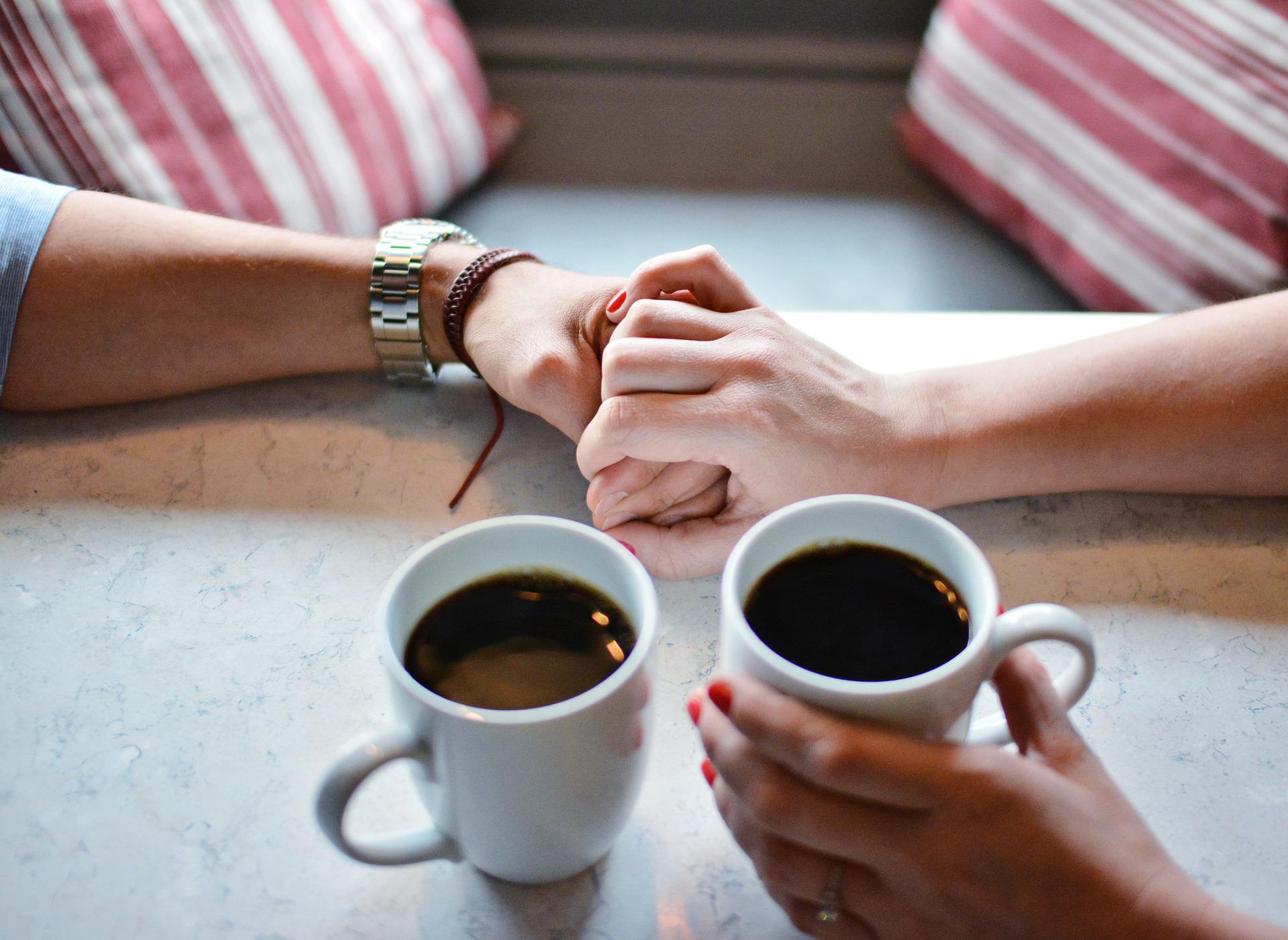 Zwei Personen, dessen Hände sich berühren. Auf dem Tisch stehen zwei Tassen Kaffee.