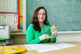 Frau sitzt an einem Pult in der Schule und hält Pokal hoch.
