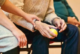Man sieht die Hand eines älteren Mannes mit einem Ball. Darüber liegt die Hand einer anderen Person