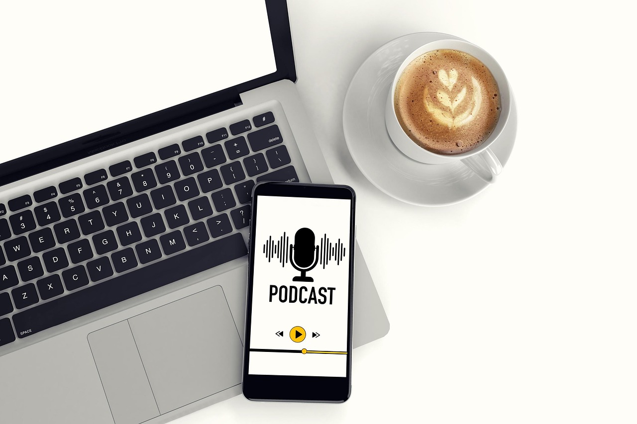Man sieht ein Handy, auf dem ein Podcast läuft auf einem Laptop, daneben eine Tasse Kaffee