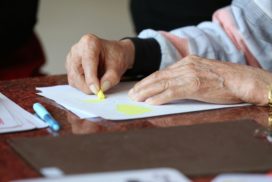 Man sieht die Hände einer älteren Person, die mit einem gelben Stift etwas auf ein Papier malen