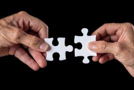 Man sieht rechts und links je eine Hand einer älteren Person mit einem Puzzleteil in der Hand, diese sich in der Mitte treffen