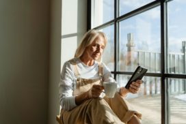 man sieht eine ältere Frau in einem Fenster sitzen mit einer Tasse in der einen und einem Handy in der anderen Hand