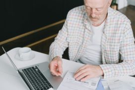 Man sieht einen älteren Mann vor einem Laptop und weiteren analogen Dokumenten an einem Schreibtisch sitzen
