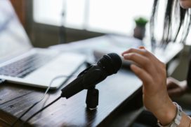 man sieht eine Hand über einem Mikrofon, das Foto suggeriert die Aufnahme eines Podcasts