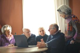 Man sieht eine Gruppe von Senior:innen miteinander sprechend vor einem aufgeklappten Laptop