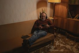 Man sieht einen älteren Mann auf einem Sofa sitzend mit Kopfhörern auf den Ohren