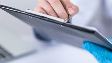 Auf einem Notizbrett wird etwas notiert. Eine Hand trägt einen Schutzhandschuh.