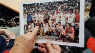 Zwei Hände halten ein älteres Mannschaftsbild des 1. FC Köln. Die linke Hand zeigt mit dem Finger auf einen Spieler.