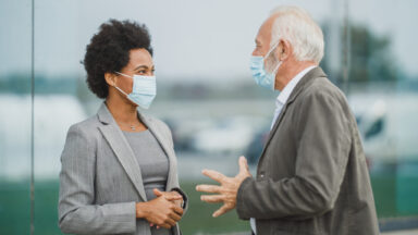 Ein Senior und eine junge Frau unterhalten sich. Sie tragen alltagsmasken und gestikulieren mit ihren Händen.
