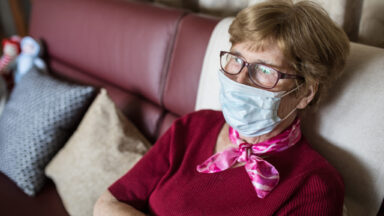 Eine Seniorin sitzt mit Maske auf ihrem Sofa.