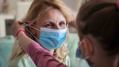 Eine Frau unterstütz eine Angehörige beim Anziehen eines Mundschutzes.