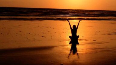 Eine Person macht Yoga am Strand mit dem Blick in Richtung Sonnenuntergang.