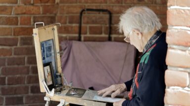 Ein Mann sitzt draußen an einem kleinen Tisch und malt ein Bild.