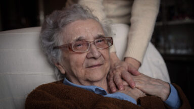 Eine Seniorin sitzt auf einem Sessel und hält die Hand einer jüngeren Frau.