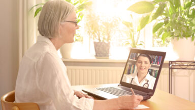 Eine Seniorin sitzt am Tisch in ihrer Wohnung und nimmt an einer Onlinekonferenz teil.