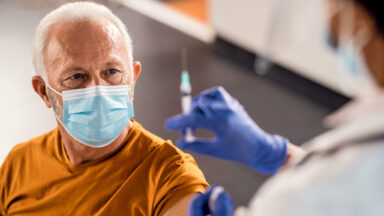 Ein Senior bekommt eine Impfung. Alle tragen medizinische Atemschutzmasken.