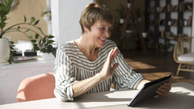 Eine Frau sitzt an einem Tisch und nutzt ein Tablet für einen Videoanruf.. Sie winkt in die Kamera.