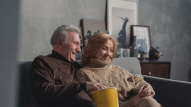 Zwei Senior*innen sitzen auf einer Couch und essen Popcorn. Sie schauen einen FIlm.