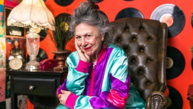 Ein Seniorin sitzt auf einem Sessel vor einer Wand mit vielen Schallplatten.