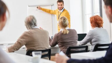 Foto aus einem Seminar. Der Dozent zeigt auf das Flipchart und eine Teilnehmering stellt eine Frage.
