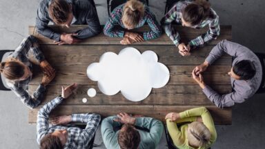 Achte Personen sitzen um einen Tisch und besprechen sich. In der Mitte des Tisches ist eine symbolische Denkblase.