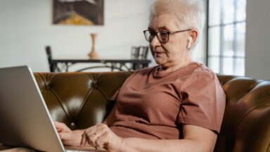 Eine Seniorin sitzt auf der Couch und nimmt über ihren Laptop an einem Online-Seminar teil.